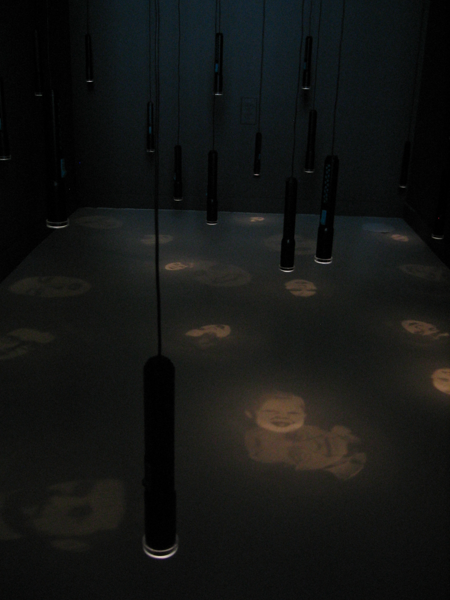 Flashlights projecting faces hang down from a dark gallery ceiling