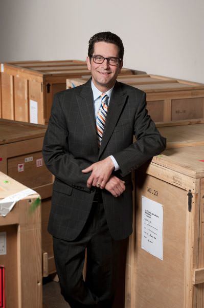 Person with glasses poses next to a shipping box in a suit