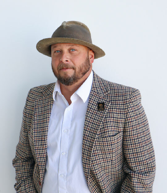 A white man with blue eyes and a brown beard is wearing a wide-brim hat, a checkered sport coat and white button up.