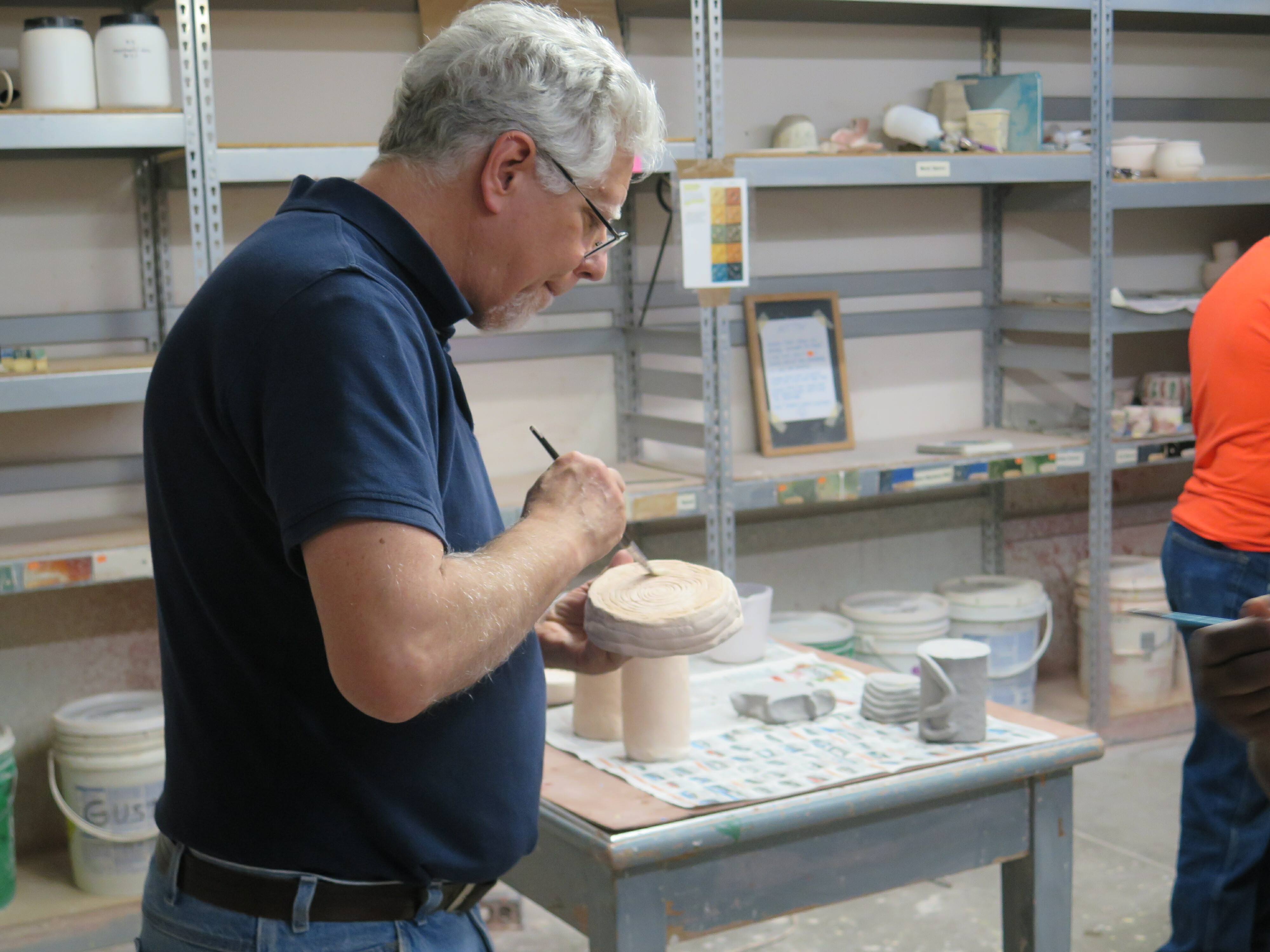 A man in a dark blue shirt is working on a ceramic piece, a tool is in his hand as he is working on the bottom. Behind him are shelves with difference ceramic pieces scattered.