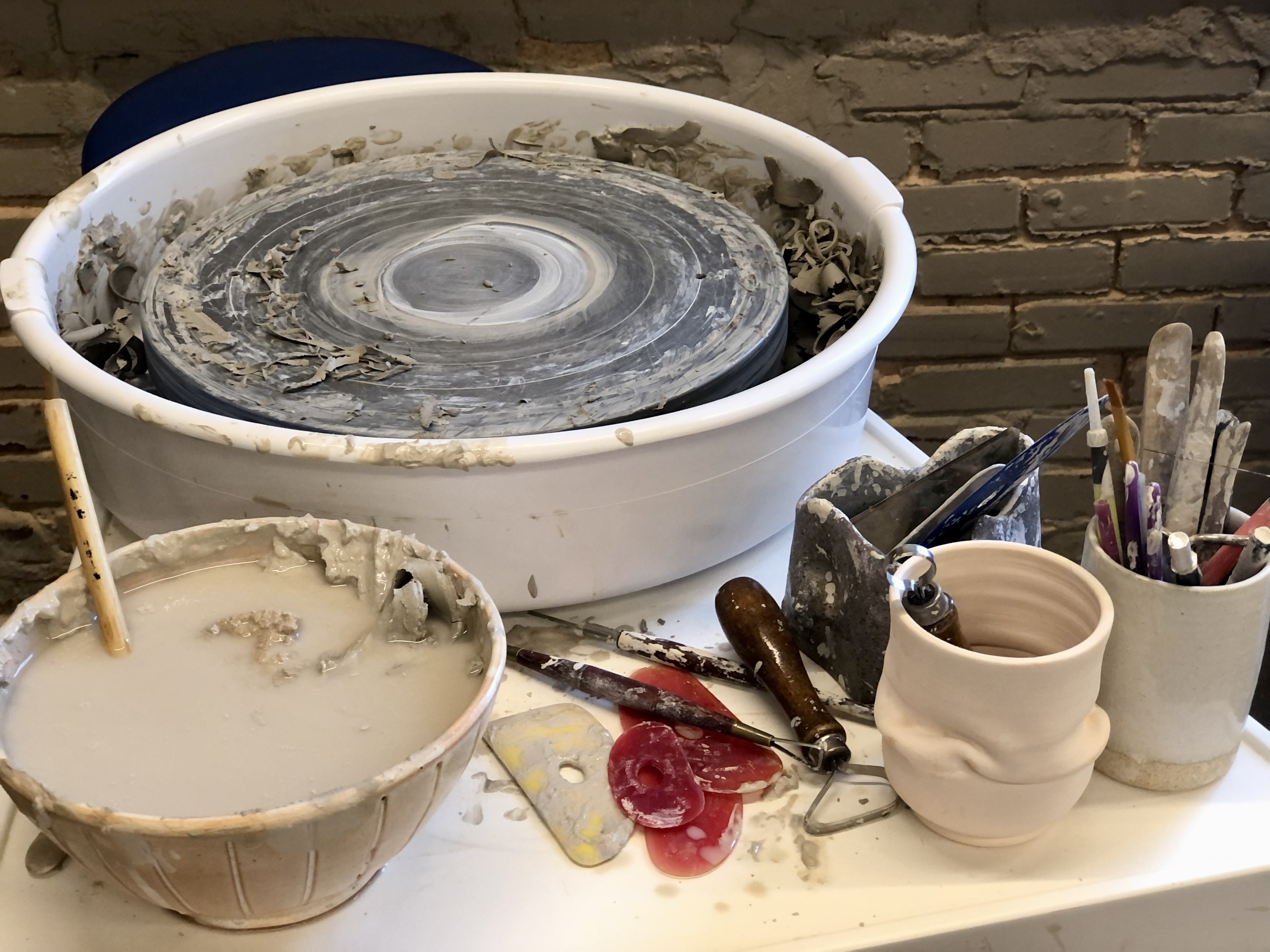 A dirty throwing wheel is covered in pieces of clay. A bowl with murky water sits in front, along with two clay vessels, both beige, one holding used brushes and tools.