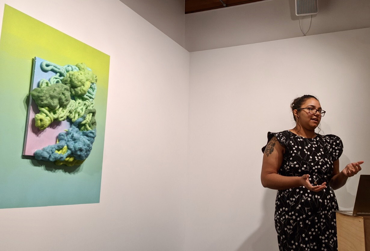 The artist stands at a podium in a black and white polka dot dress with thin-rfamed glasses on and her hair in a bun. She is talking. Behind her is a bright green and blue piece with textured fibers and materials bubbling off the surface