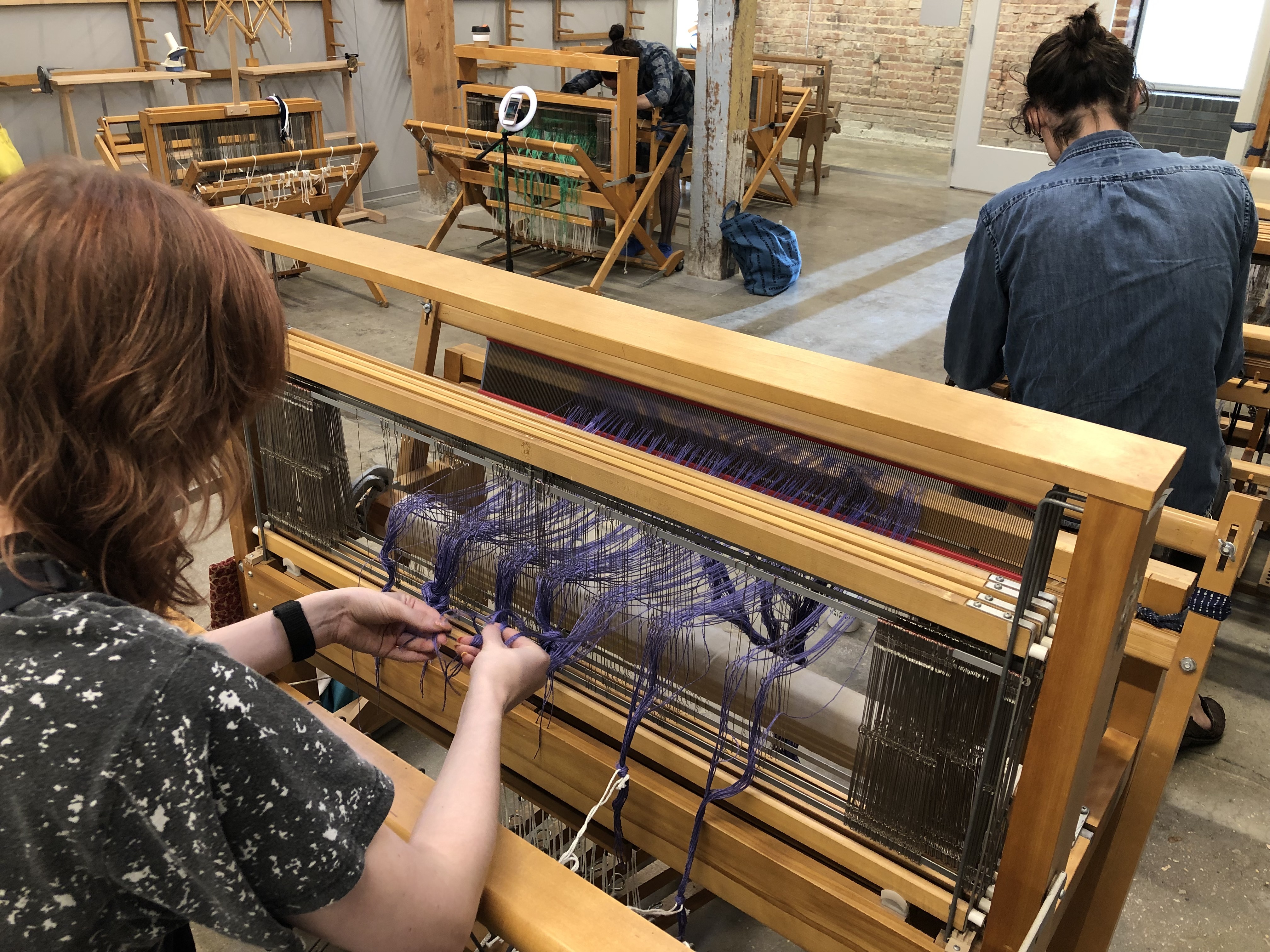 There are three people working on three, separate looms. Two have their backs to the camera and we can see one is working with purple thread as they pull it through the loom. In the back, the third person is facing us, bent over the loom.