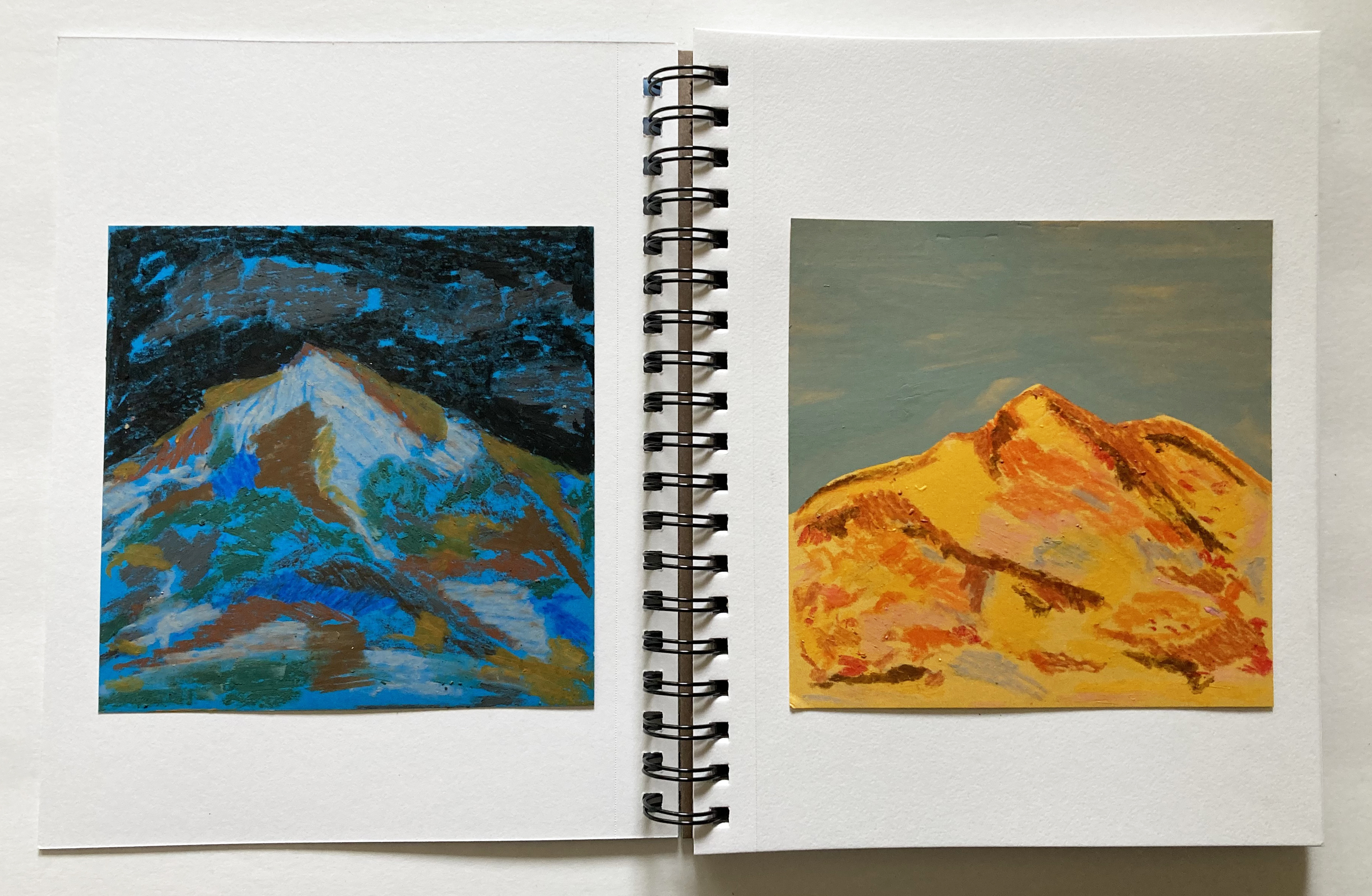 A spiral-bound journal is open. The pages are white with images. On the left, blyes, browns and greens create a night scene of a mountain. The left, yellows and oranges create a daytime scene.