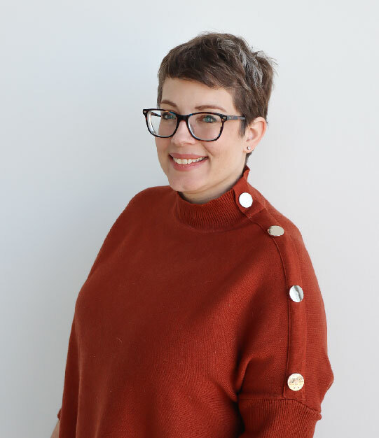 A white woman with short brown hair wearing an auburn colored sweater and thick framed glasses stands in front of a white wall smiling