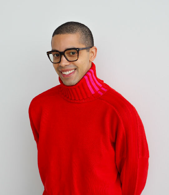 A Black person with short buzzed hair, a bright red turtleneck sweater and thick-rimmed glasses smiles at the camera