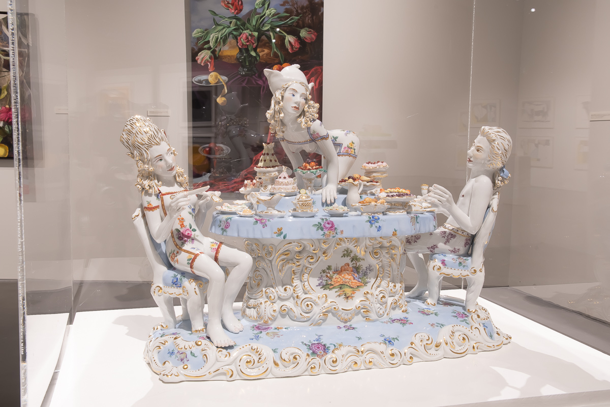 A porcelain figurine depicting a woman on all fours on a table with food. A man and another woman sit at either end of the table. A still life featuring fruit is in the background.