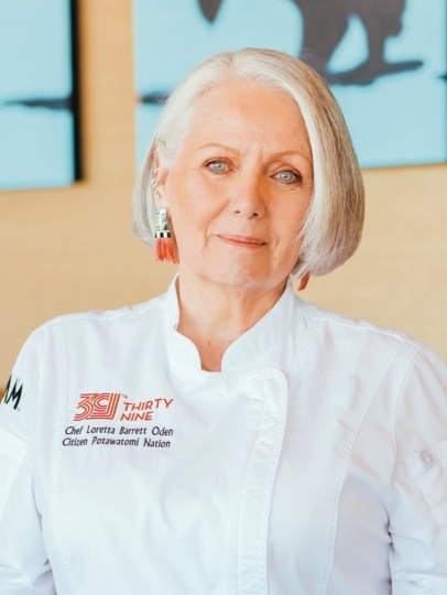 A white-haired person wearing a chef's coat and red earrings