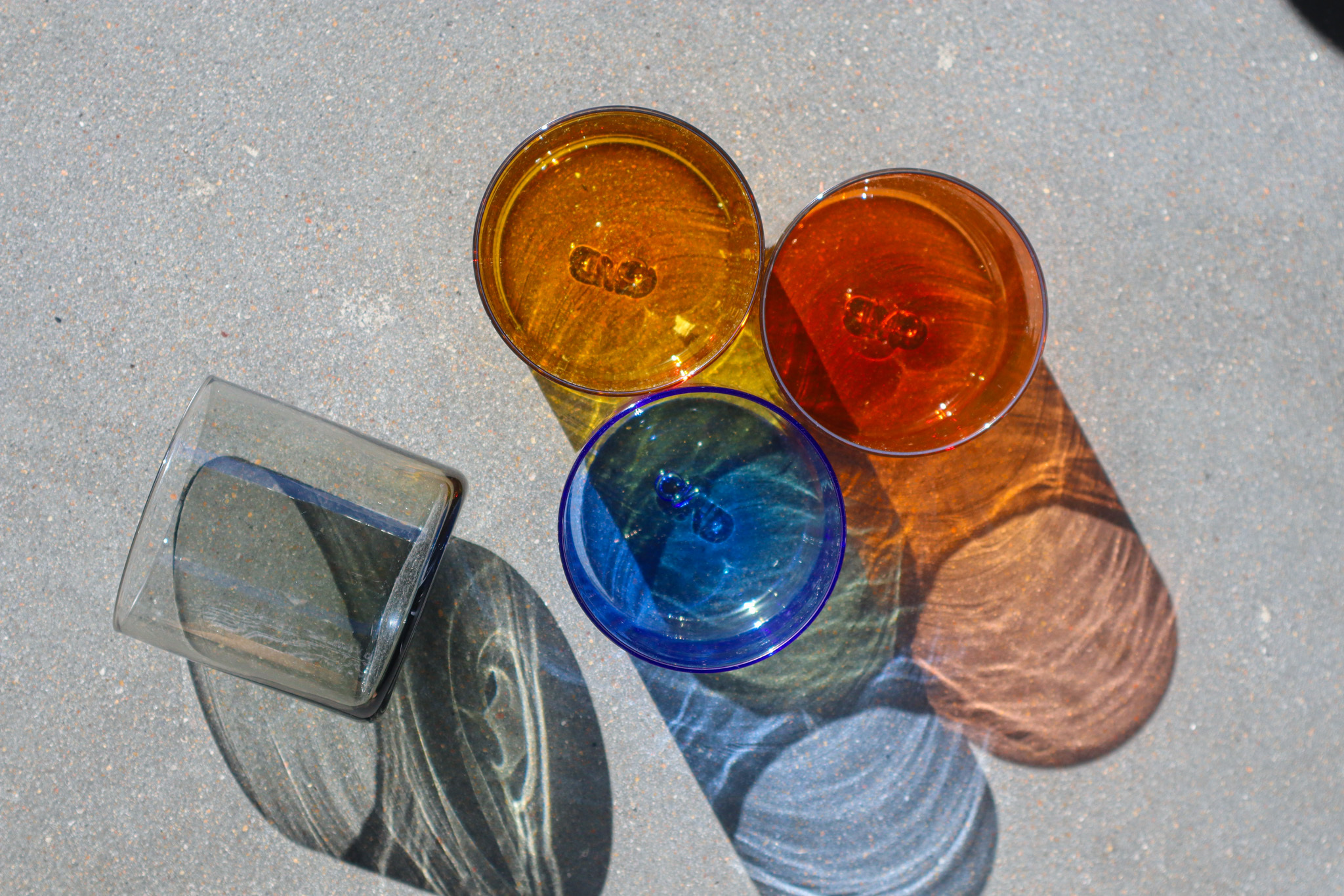 Four glass cups sit on cement. Three are grouped in a triangle while one lays on its side. The group of three are orange, red and blue colors, and the single glass is black.