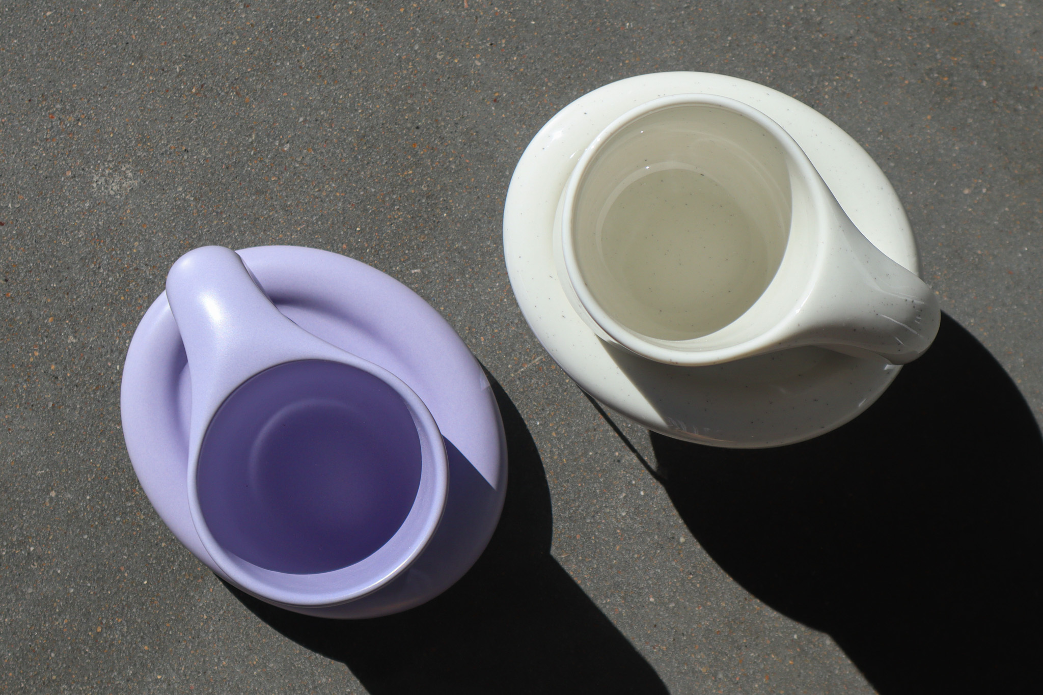 Two mugs with matching saucers sit on a cement surface. On the left is a lavender colored mug, and on the right a white mug with black flecks.