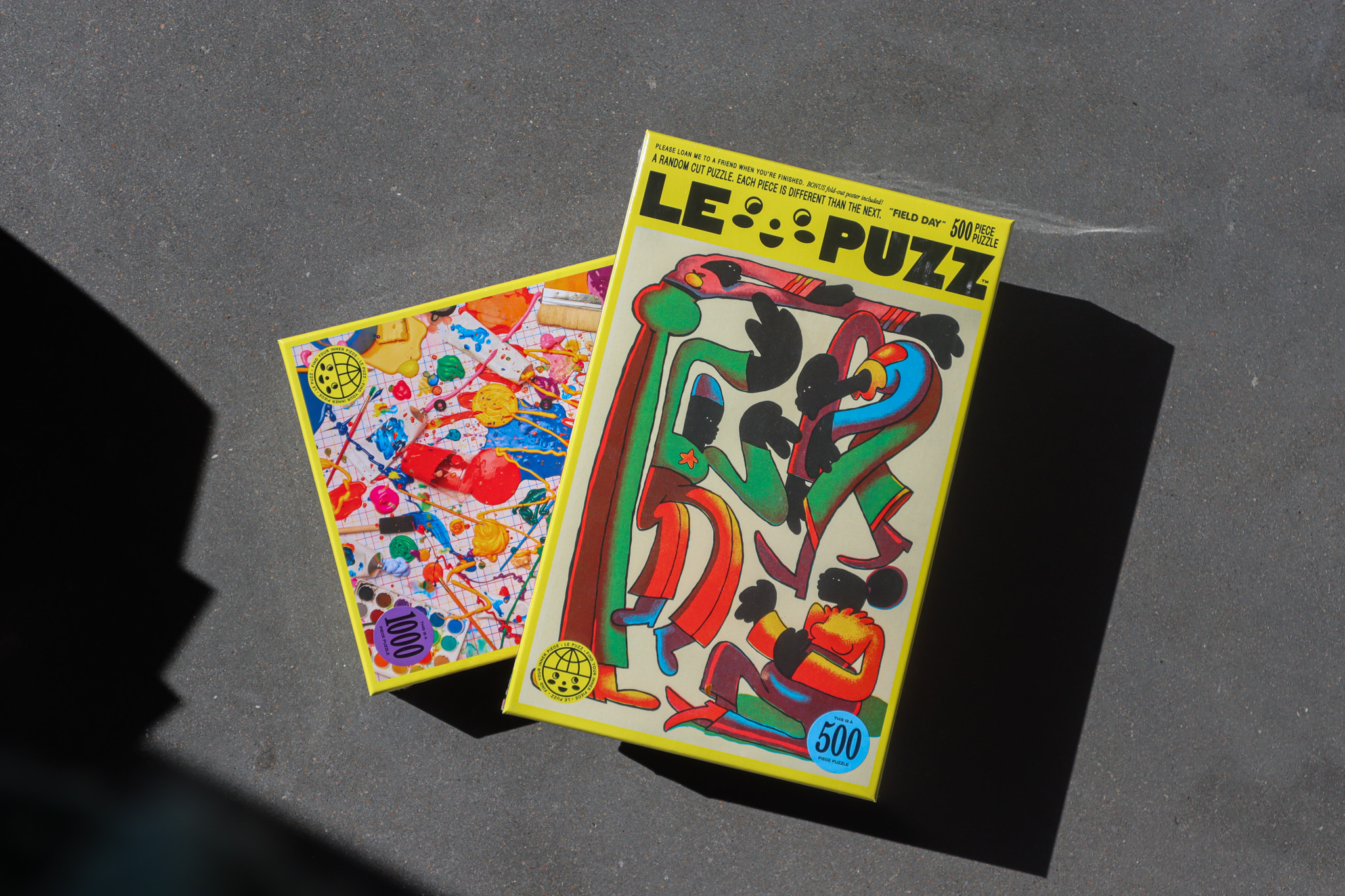Two puzzle boxes sit stacked on top of each other against a cement surface. They are filled with vibrant colors and abstract shapes, bordered in bright yellow with the words LE PUZZ written in black block text at the top.