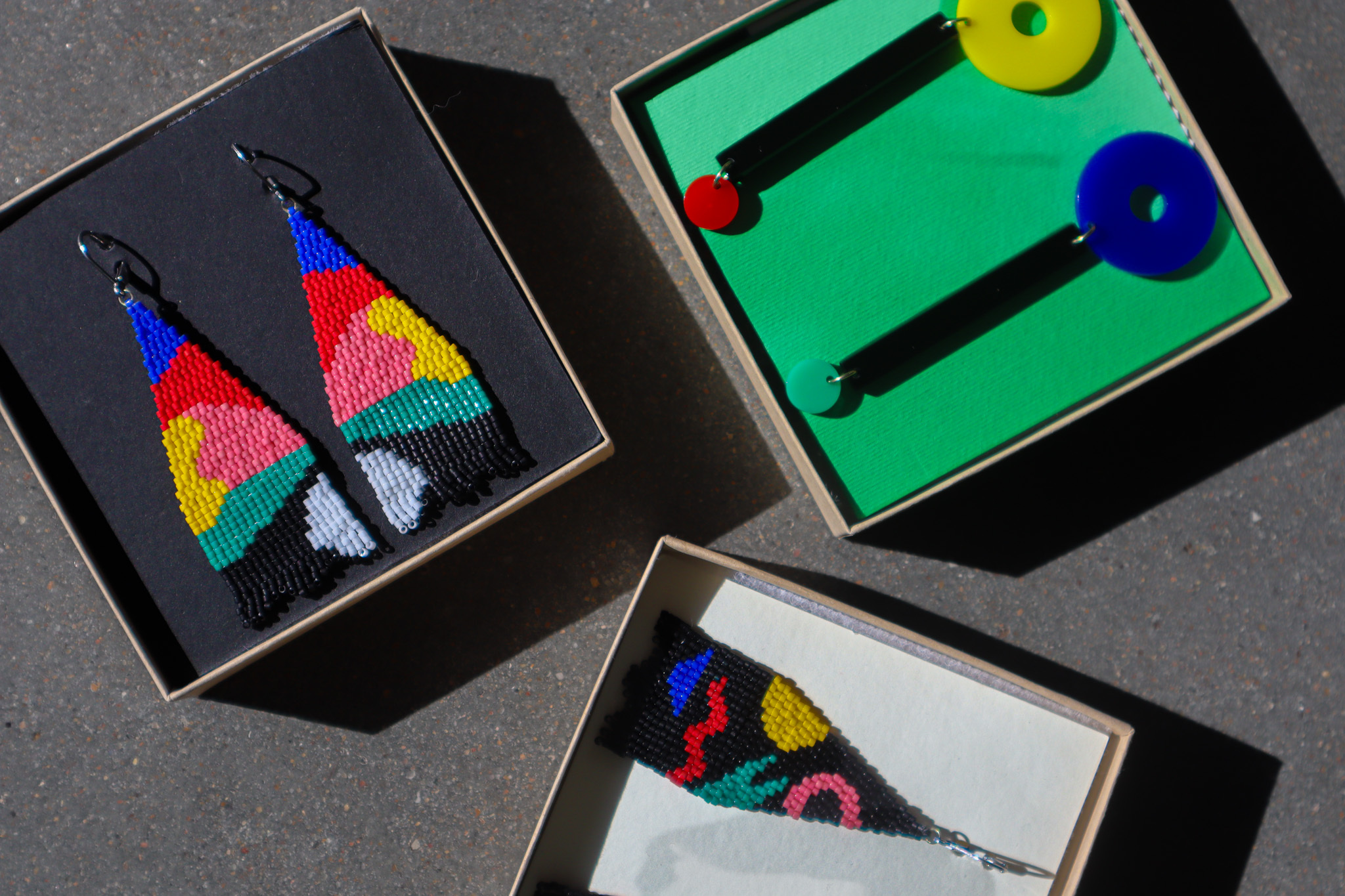 Three pairs of earrings in wooden boxes sit on a cement surface. Two are out of frame; the one pair in frame is a dangling set of earrings on a black background. They are abstract shaped colors of yellow, pink, green, blue, black and white