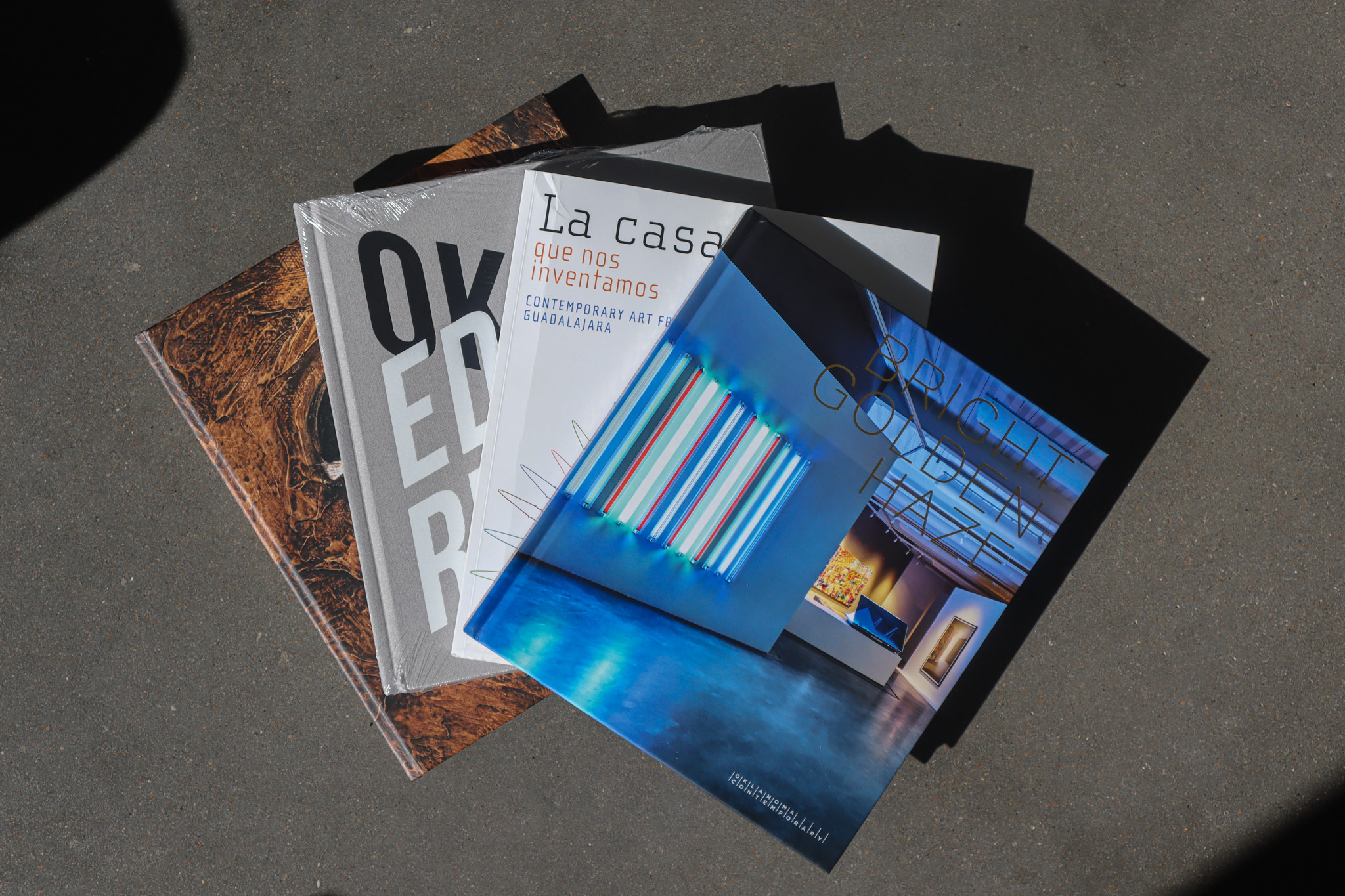 Four exhibition catalogs lay on a cement surface. They are stacked on top of each other, fanned out. The top book shows a blue hued installation with the words BRIGHT GOLDEN HAZE written on the cover in gold