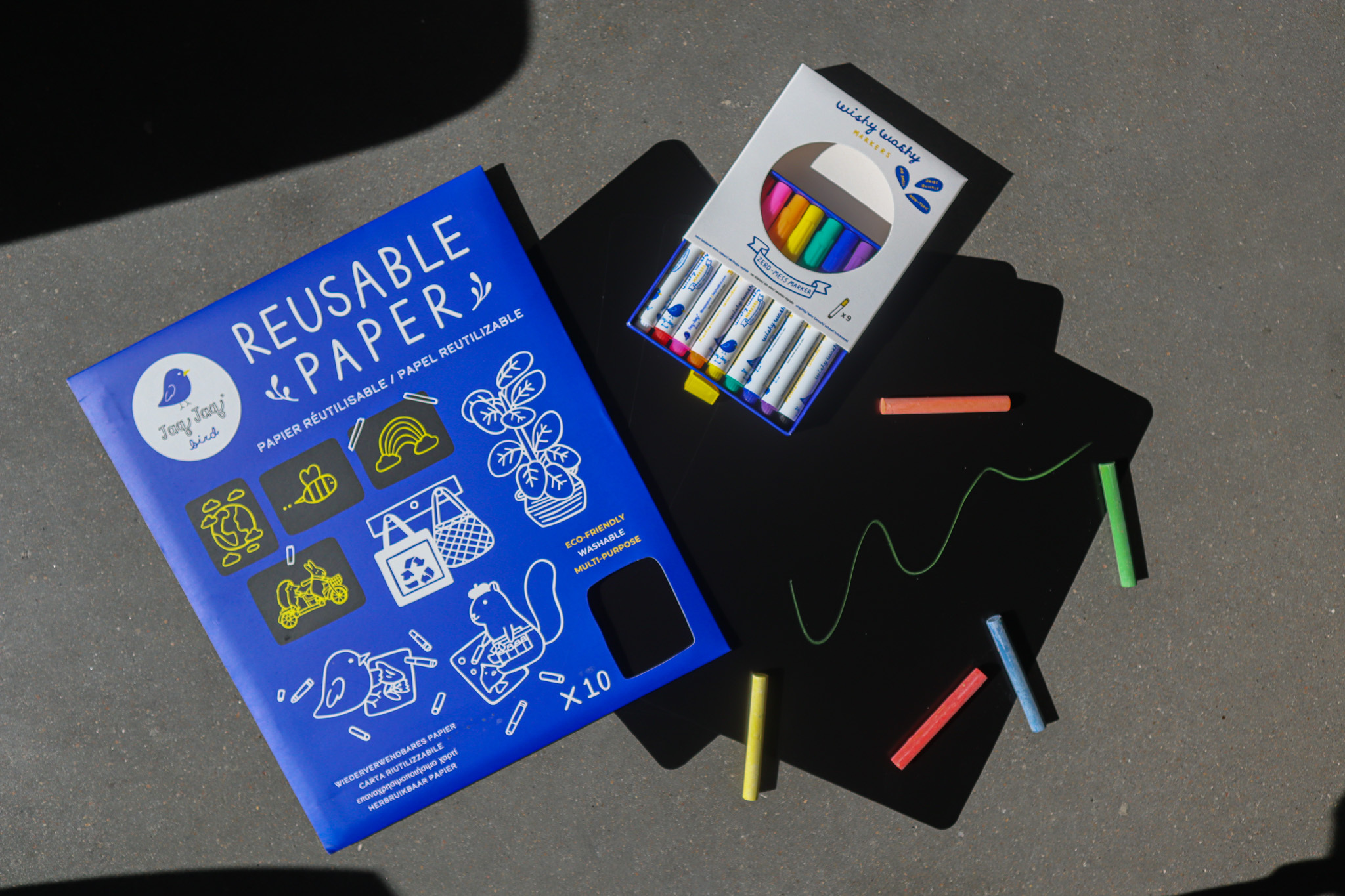 Art-making tools lay on a cement surface. On the left is a blue envelope with yellow and white doodles on the front and text that reads REUSABLE PAPER. To the right is black sheets, a box of markers and scattered crayon-like coloring sticks.