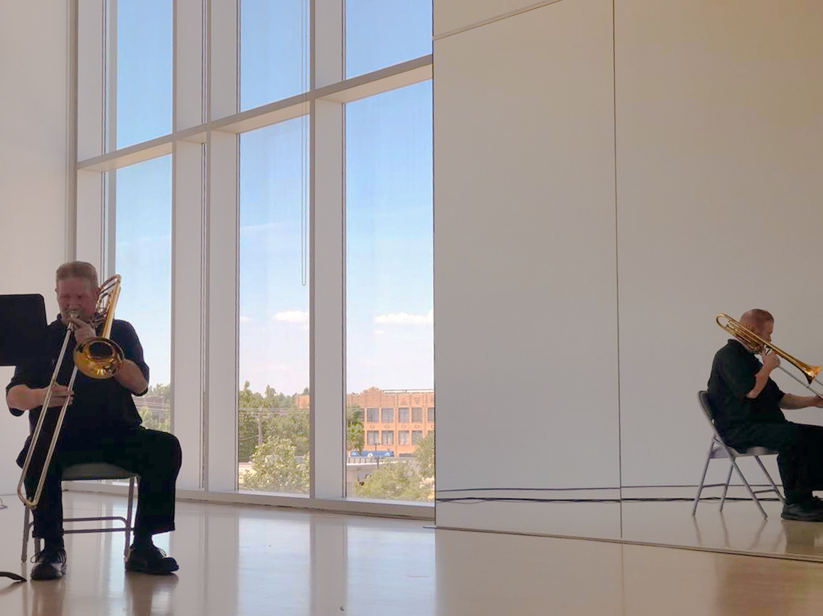 A man dressed in all black is playing the trombone, sitting down and facing the camera. Behind him is a floor-to-ceiling window. To his left, a floor-to-ceiling mirror reflects his image.