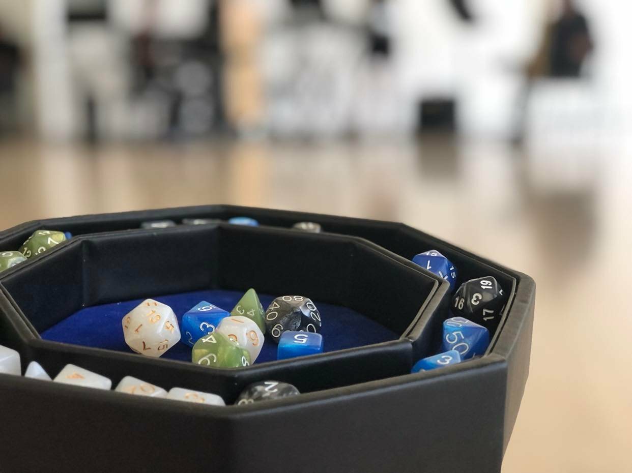 Grey, green, blue and white multi-sided dice sit in a black case with a blue, velvet base. Out of focus, we can make out people and instruments.