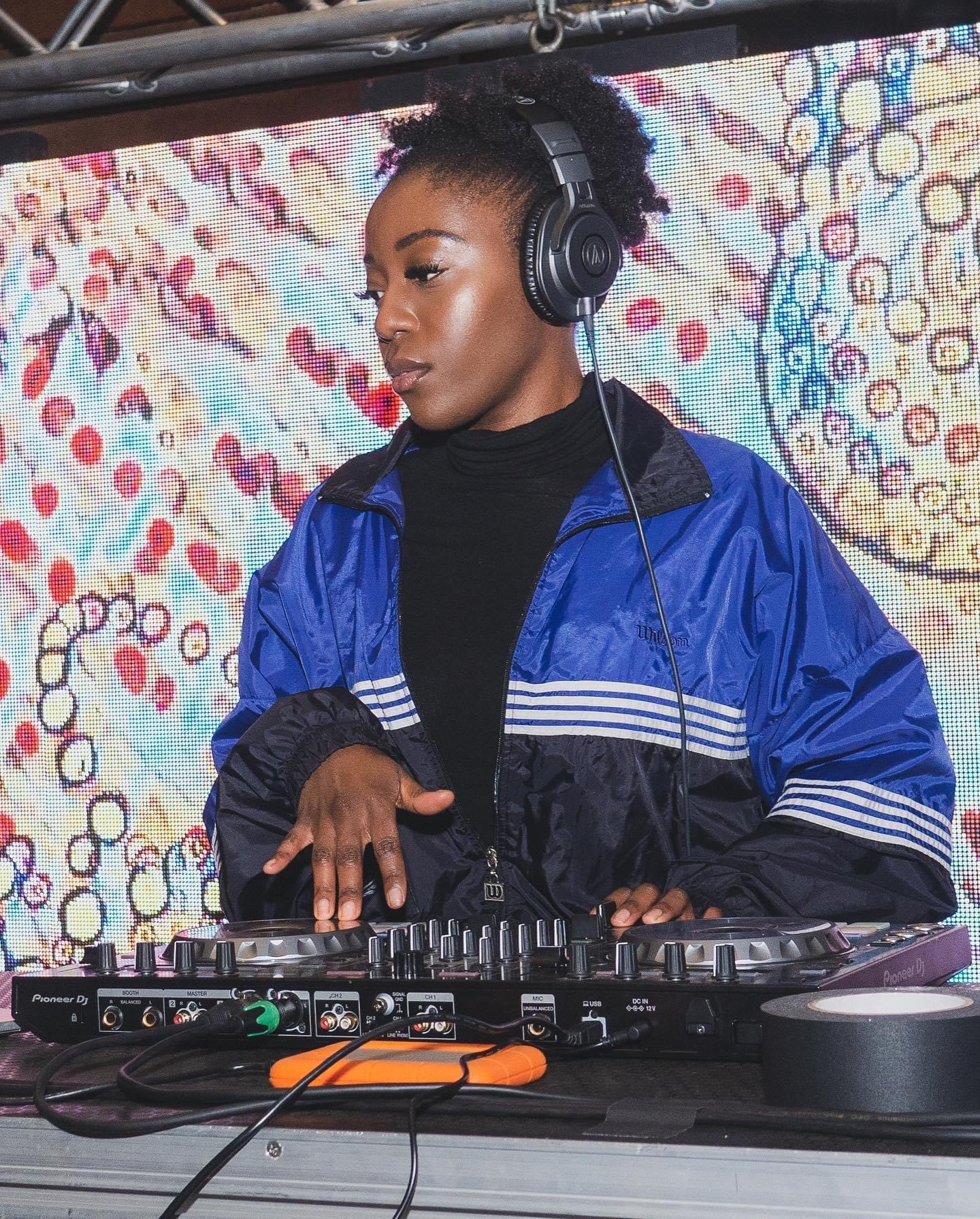 A person wearing a windbreaker and over-the-ear headphones stands at a DJ station with a colorful backdrop