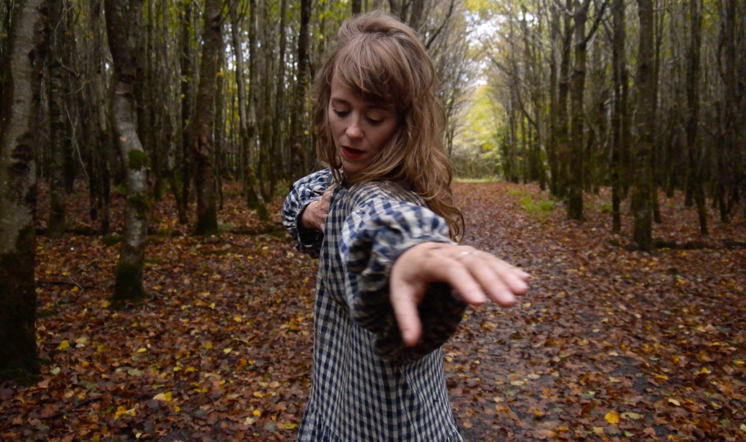 A person with dirty-blonde hair wearing a black-and-white gingham dress stands in a forest clearing with their left arm extended