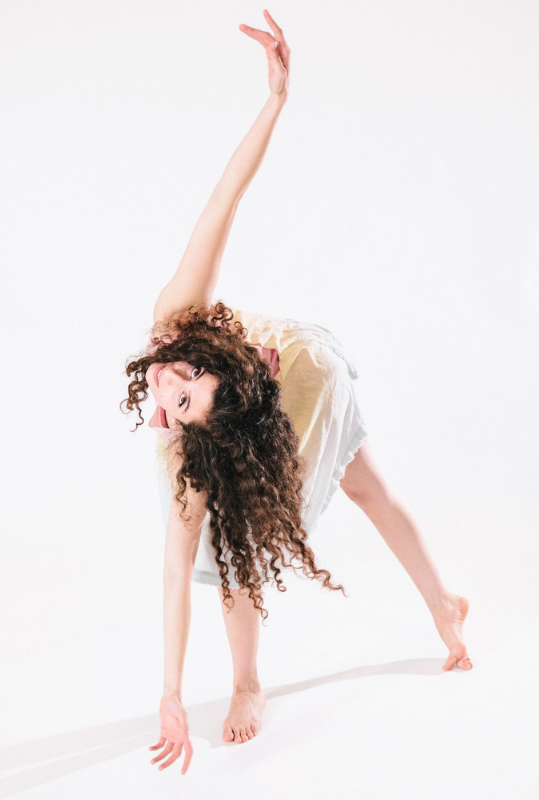 A dancer with long, curly brown hair and torso bent parallel with the ground extends one arm to the floor and the other to the ceiling against a white backdrop