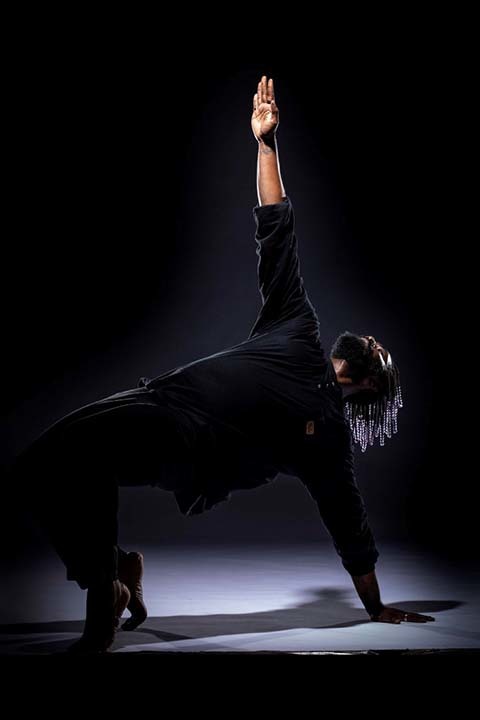 A dancer wearing all black in a dimly lit space arches backwards with one arm extended upwards