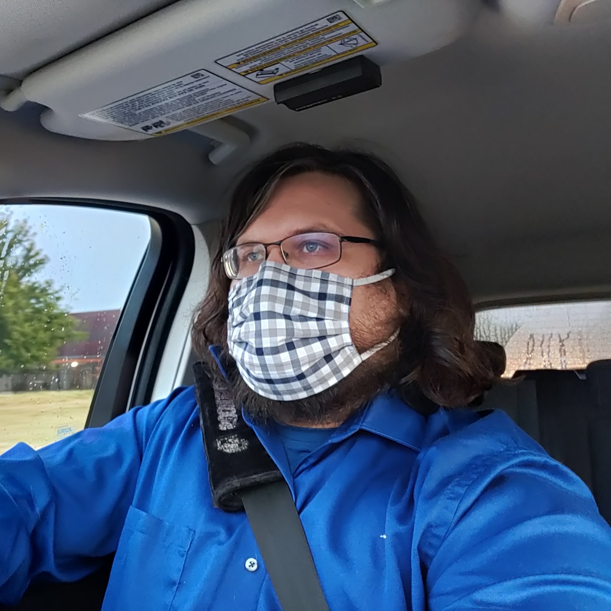 A person with shoulder-length dark hair, gingham mask, blue button-down shirt and rectangular glasses sits in the front seat of a car