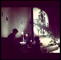 Two people sit behind a projector screen, the projector on. On the screen, shadow puppets are telling a story.