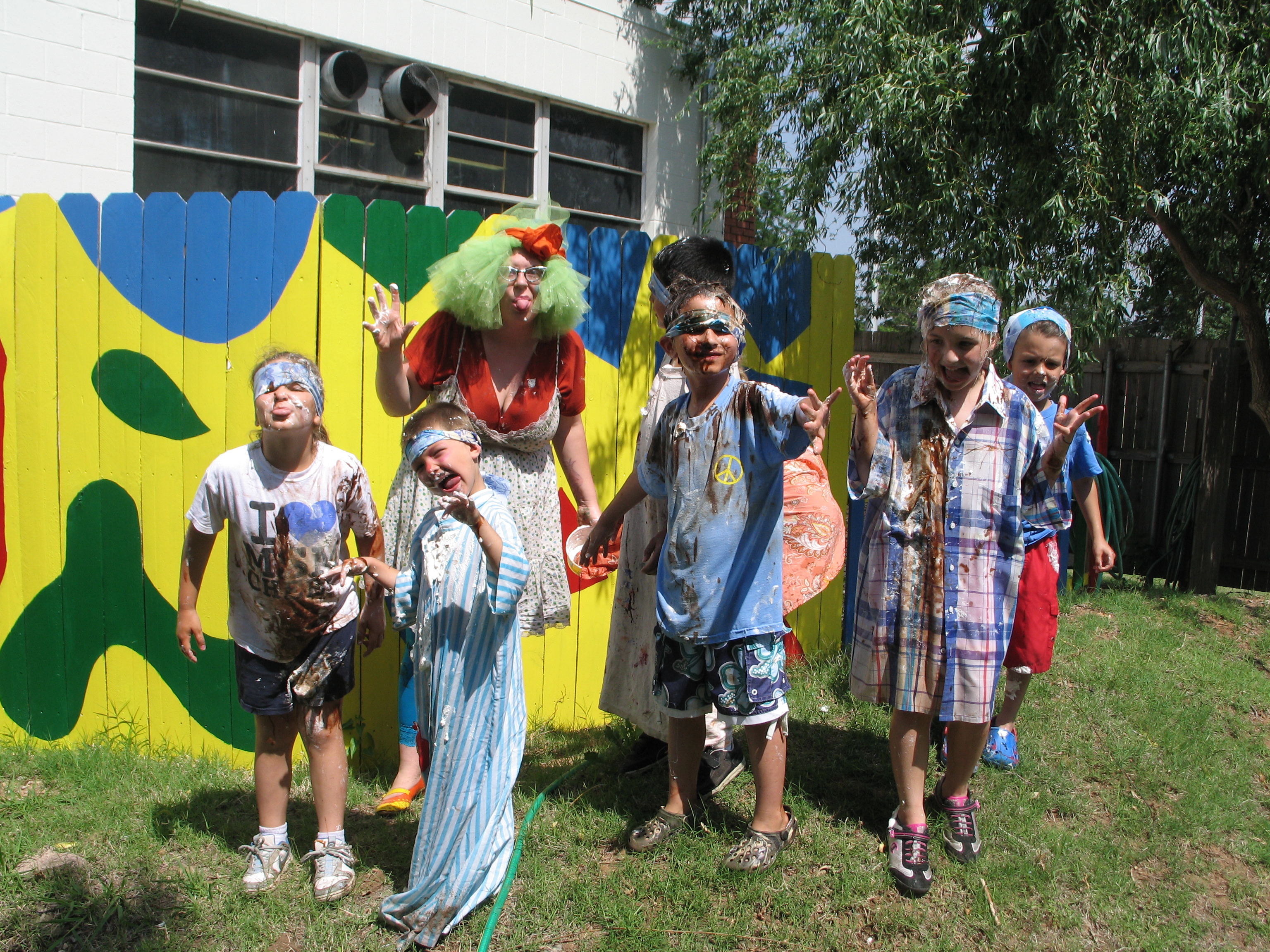 Kids stand in front of a yellow painted fence, covered in chocolate and whipped cream with bandana's on their foreheads, making silly faces. Behind them stands a person in green hair with a big red bow, sticking their tongue out.