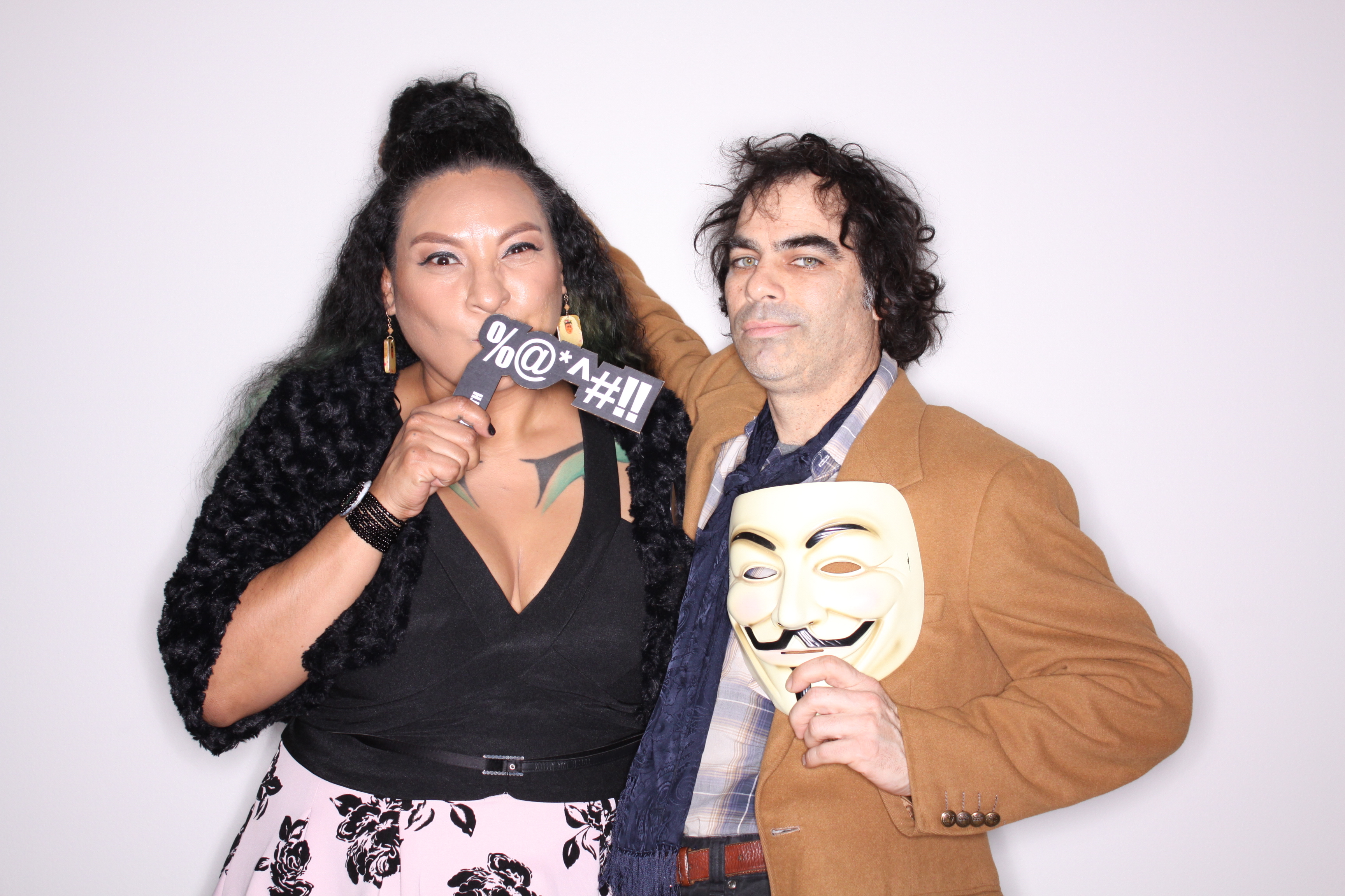 A woman in a black dress with a pink and black skirt is holding a small black sign with a %@*^#!! printed in white front of her mouth. A man in a caramel colored jacket stands next to her holding a white mask.