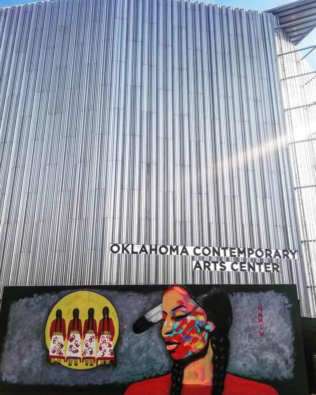 Photo of the new building with it's reflective fins, the sun shining through. You can see Oklahoma Contemporary. In front is a painted piece of an Indigenous figure right of four other figures in a yellow circle. A hashtag #MMWI in red is furthest right.
