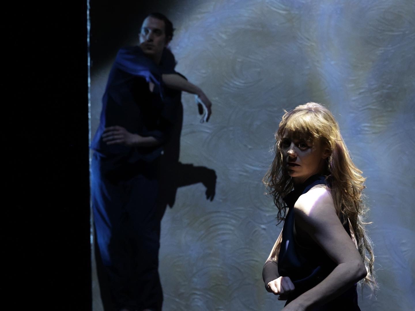 Two people are dancing. In the foreground, a woman with long blonde hair wearing a dark dress looks over her shoulder. In the background, a man wearing dark clothing stretches a hand out. Ambient blue lighting encompasses them.