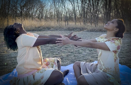 Two women have their arms outstretched toward one another, sitting on the ground outside. They are both dressed in light pastel yellows and floral.