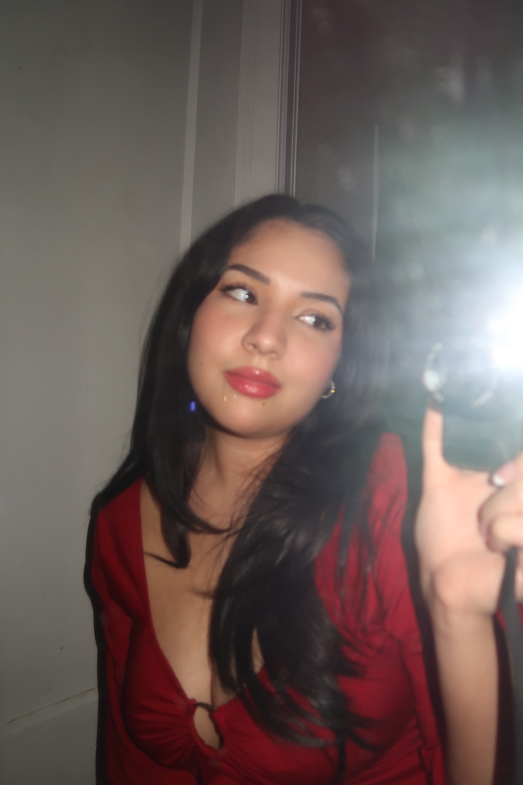 A person is taking a photo in the mirror, the camera flash shining to the right of the image. This person has long brown hair and is dressed in a red dress. Leaning forward slightly with a soft smile.