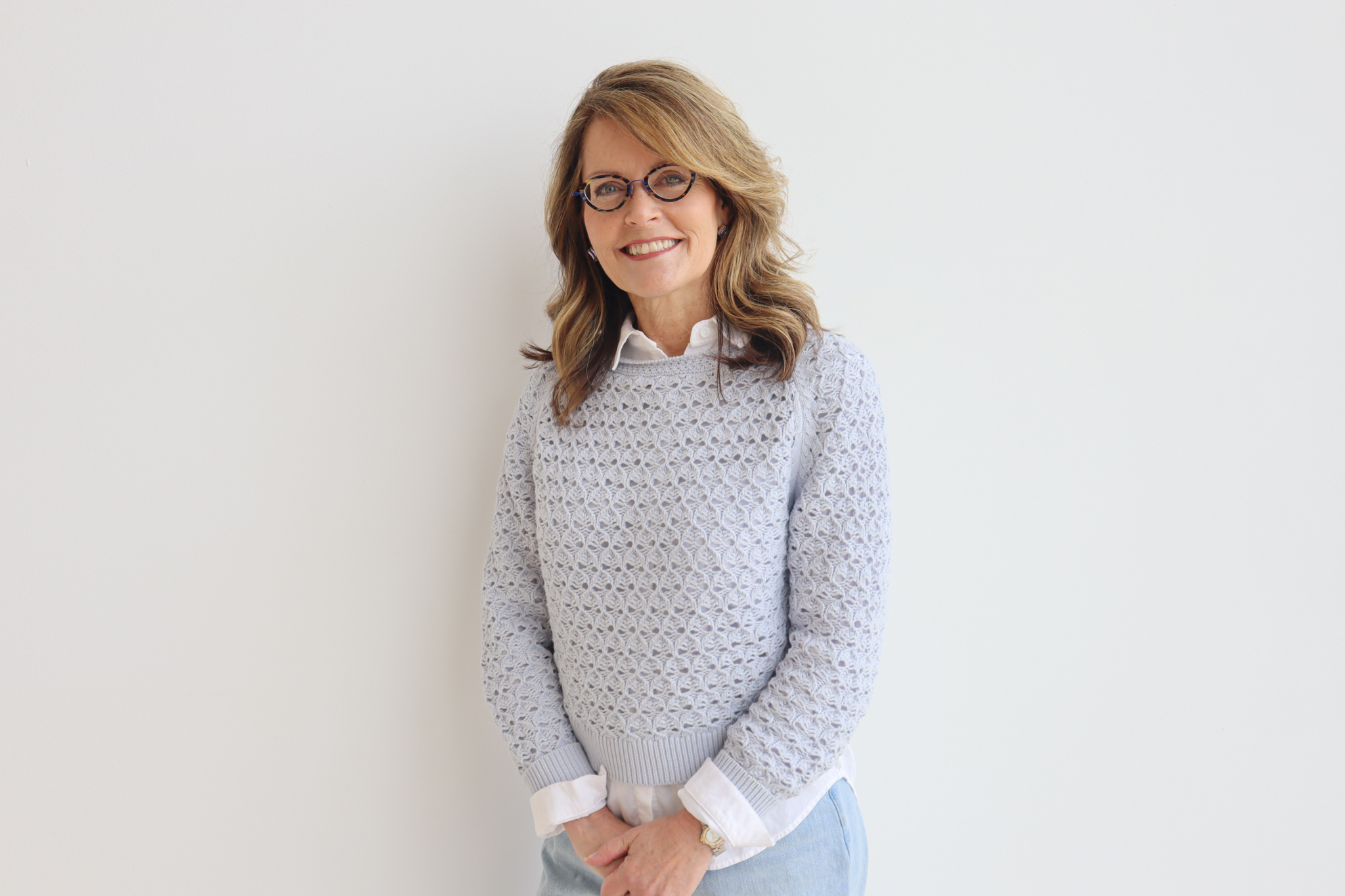 A person stands in front of a white wall. They have on a light blue sweater with white cuffs and white collar. Their hair is light brown, falling at their shoulders. They have small round glasses on and are smiling.