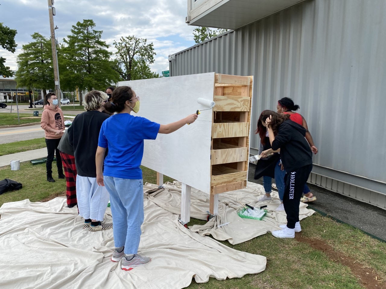 A group of teens in masks are outside. A white sheet is on the green grass underneath them as they paint a large, moveable wooden mural with white paint.