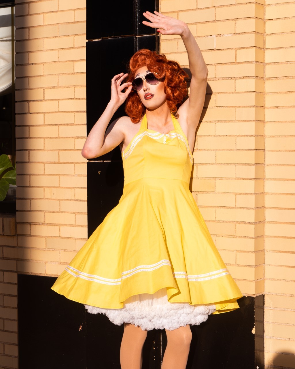 Drag queen Holly Wood Hills in a yellow, vintage cocktail dress and fiery red wig with dark sunglasses stands in the sun, back against a brick building. She is peering at the sun, one hand holding her glasses, the other stretched in front of her.
