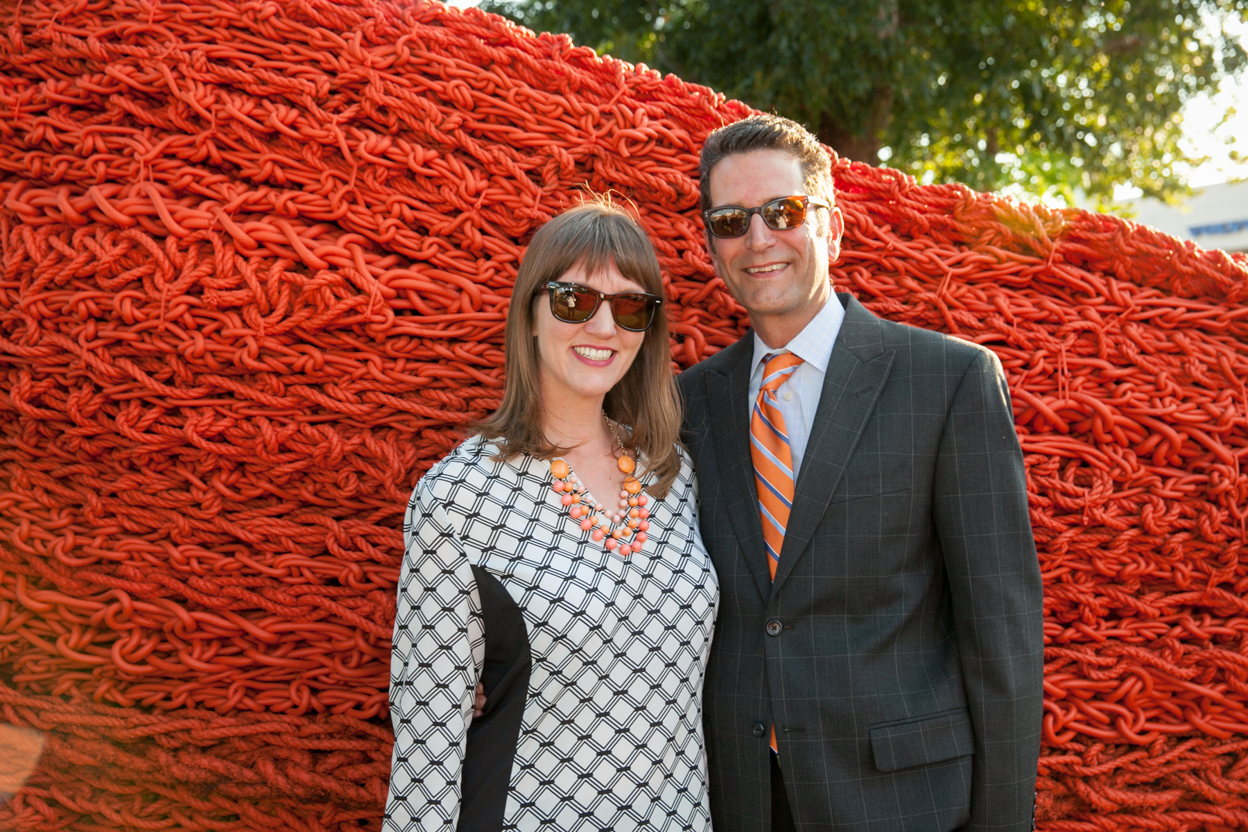 A couple smiles at the camera, dressed in a black in white patterned dress and a dark gray suit. They are standing against a wall of red lobster rope.