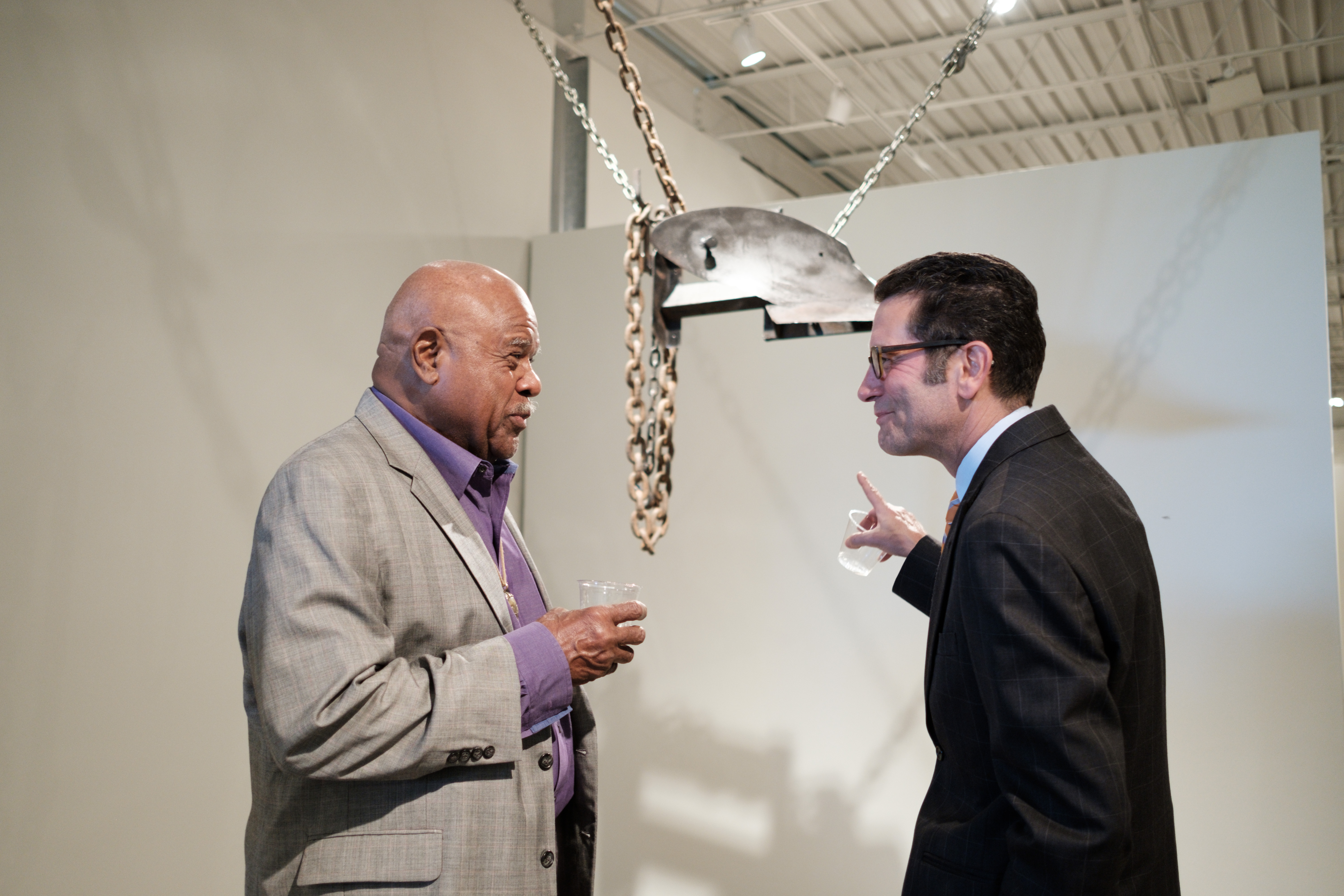 Two men are talking to each other in front of a multimedia piece of art hanging from the ceiling. They are both in suits.