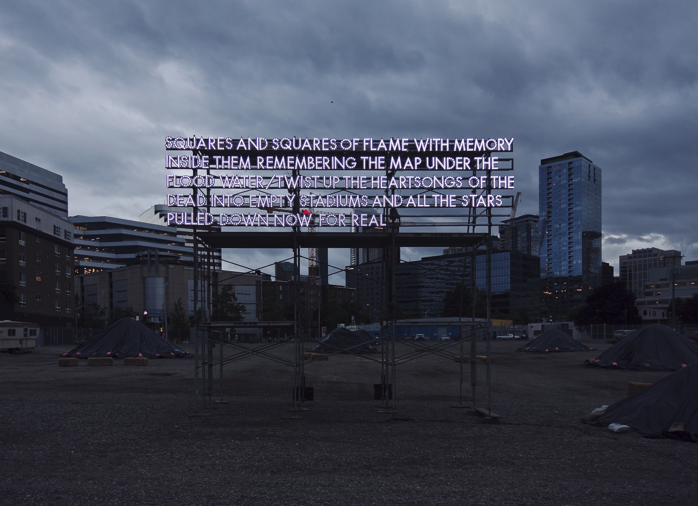 A billboard sculpture of white fluorescent lights reads “Squares and squares of flame with memory inside them remembering the map under the flood water/twist up the heart songs of the dead into empty stadiums and all the stars pulled down now for real”