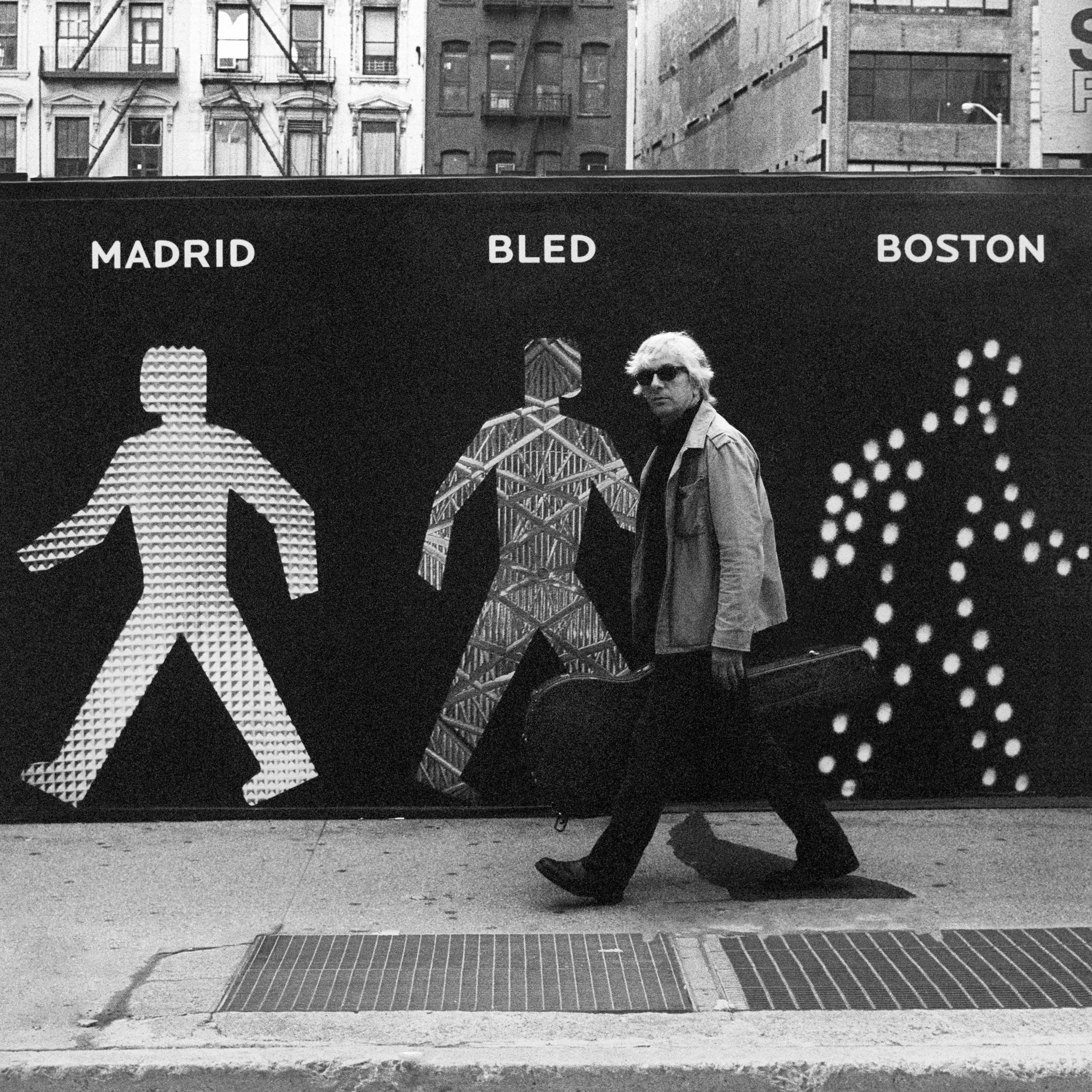 A black-and-white photo depicts a figure carrying a guitar case, against a background depicting pedestrian traffic sign outlines