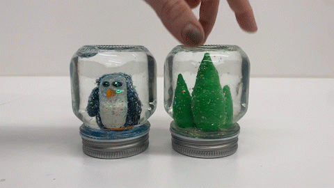 A GIF shows two small, mason-jar style snow globes. One has a little penguin inside, the other three green trees in varrying sizes. A hand picks the tree snow globe up, shakes it off camera, and puts it back down as the glitter swirls inside
