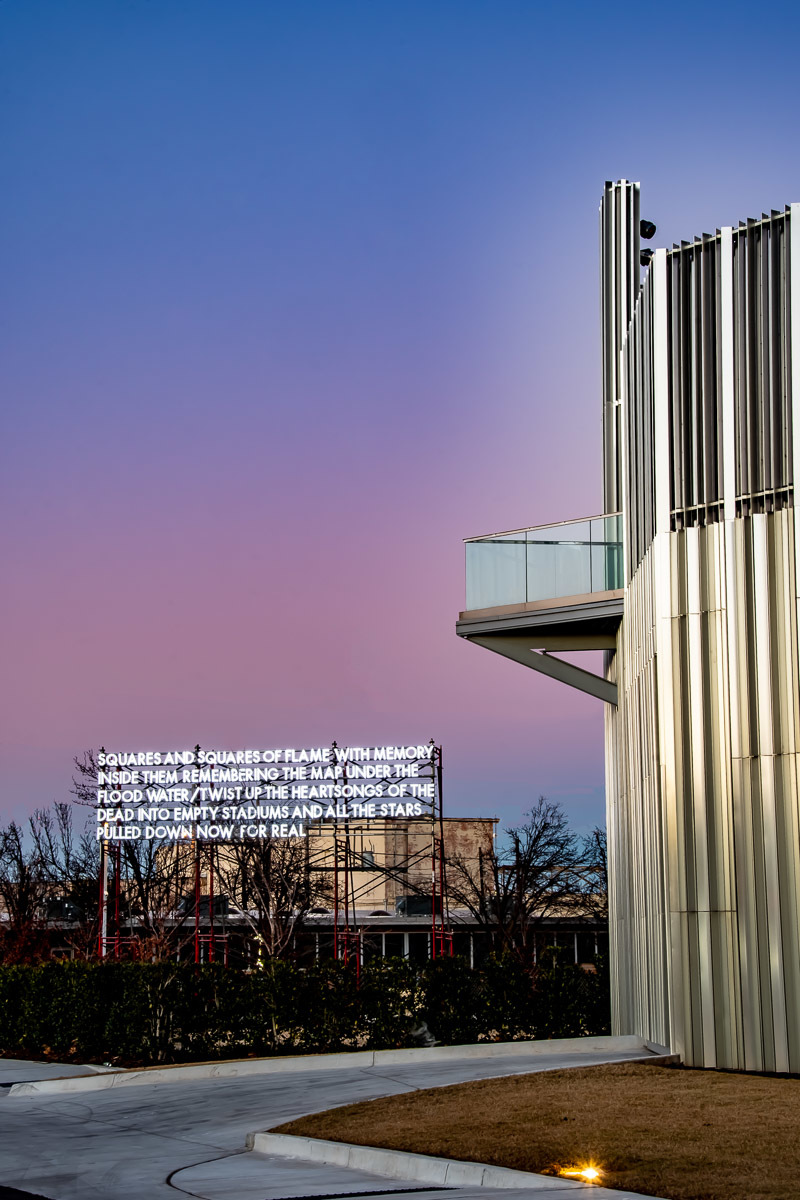 An LED billboard poem stands lit at sundown next to a silver building