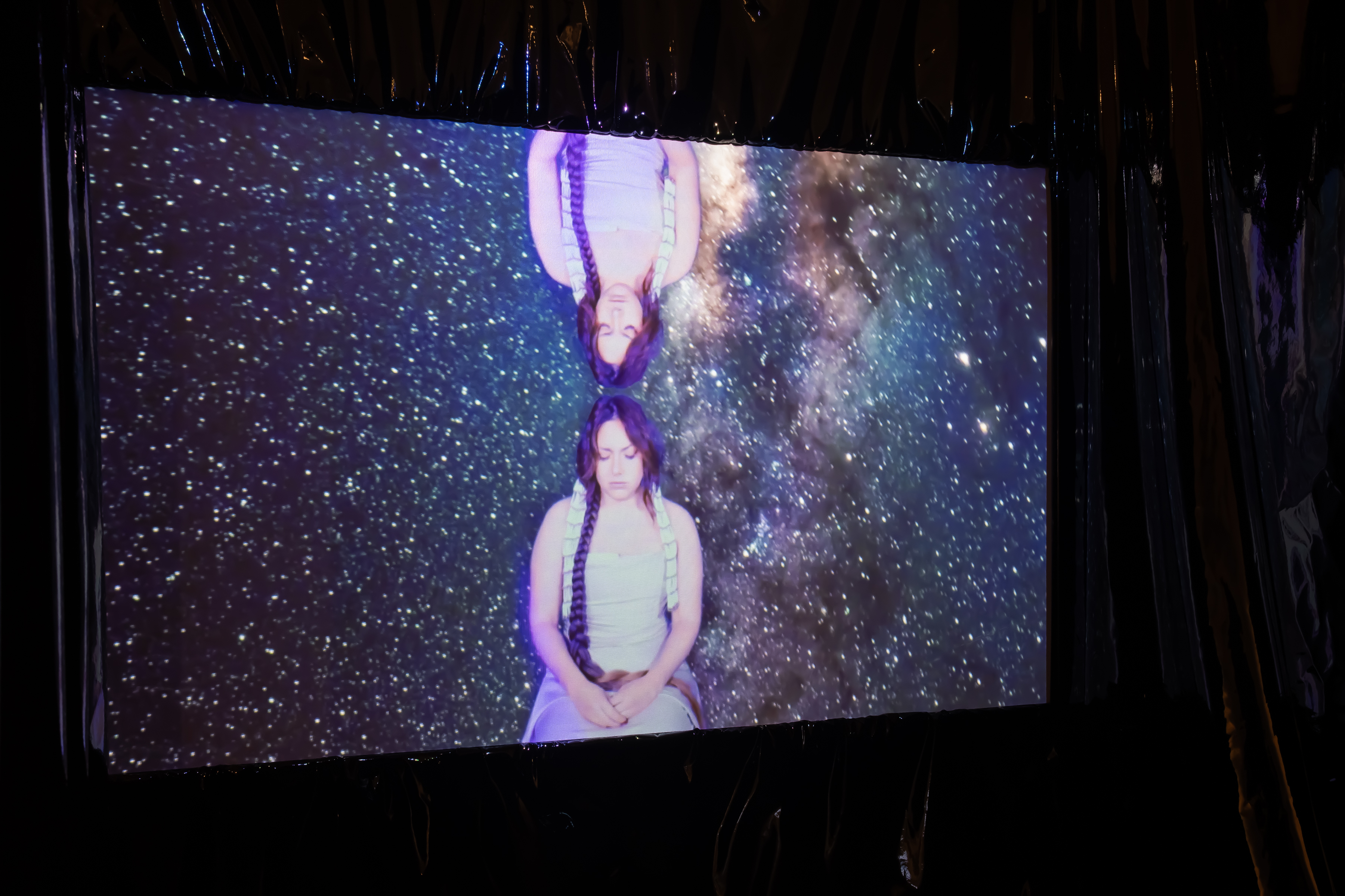 On a screen against a black wall, a person with long braided dark hair sits upright against a backdrop of constellations. A reflected image of this person is right above them.