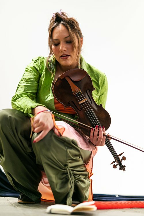 A person dressed in a green jacket and silky green pants is crouching down against a white background. They hold a dark brown violin under their chin.