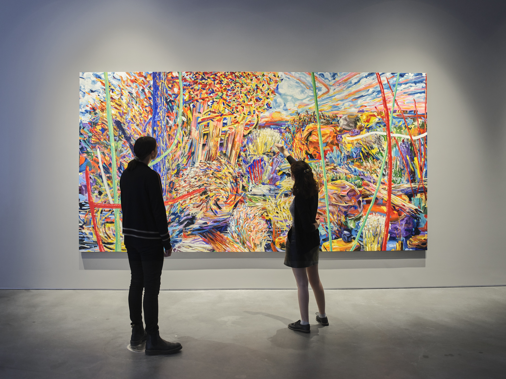 Two adults stand in front of a large, brightly colored painting of an abstract landscape