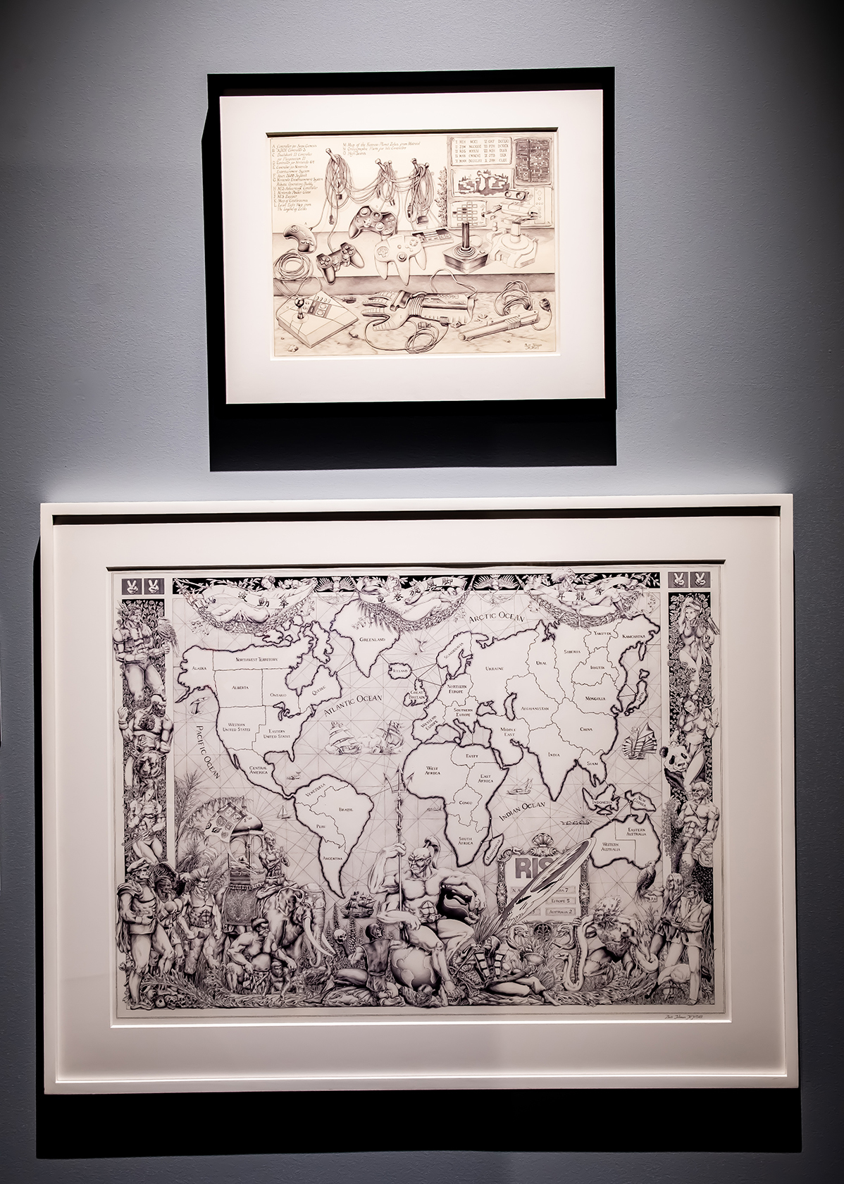 Two detailed drawings hang in frames on a gray gallery wall, one includes a map similar to a Risk board with figures at the bottom, and the other is of various video game controllers