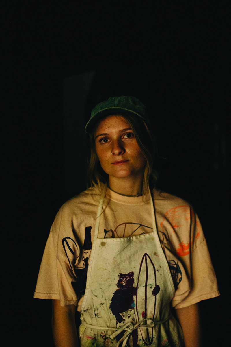 A person stands in a ballcap and a paint-splattered apron