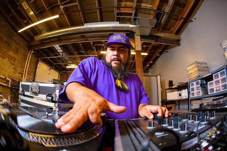 A bearded DJ with a purple hat and shirt stands at a turntable