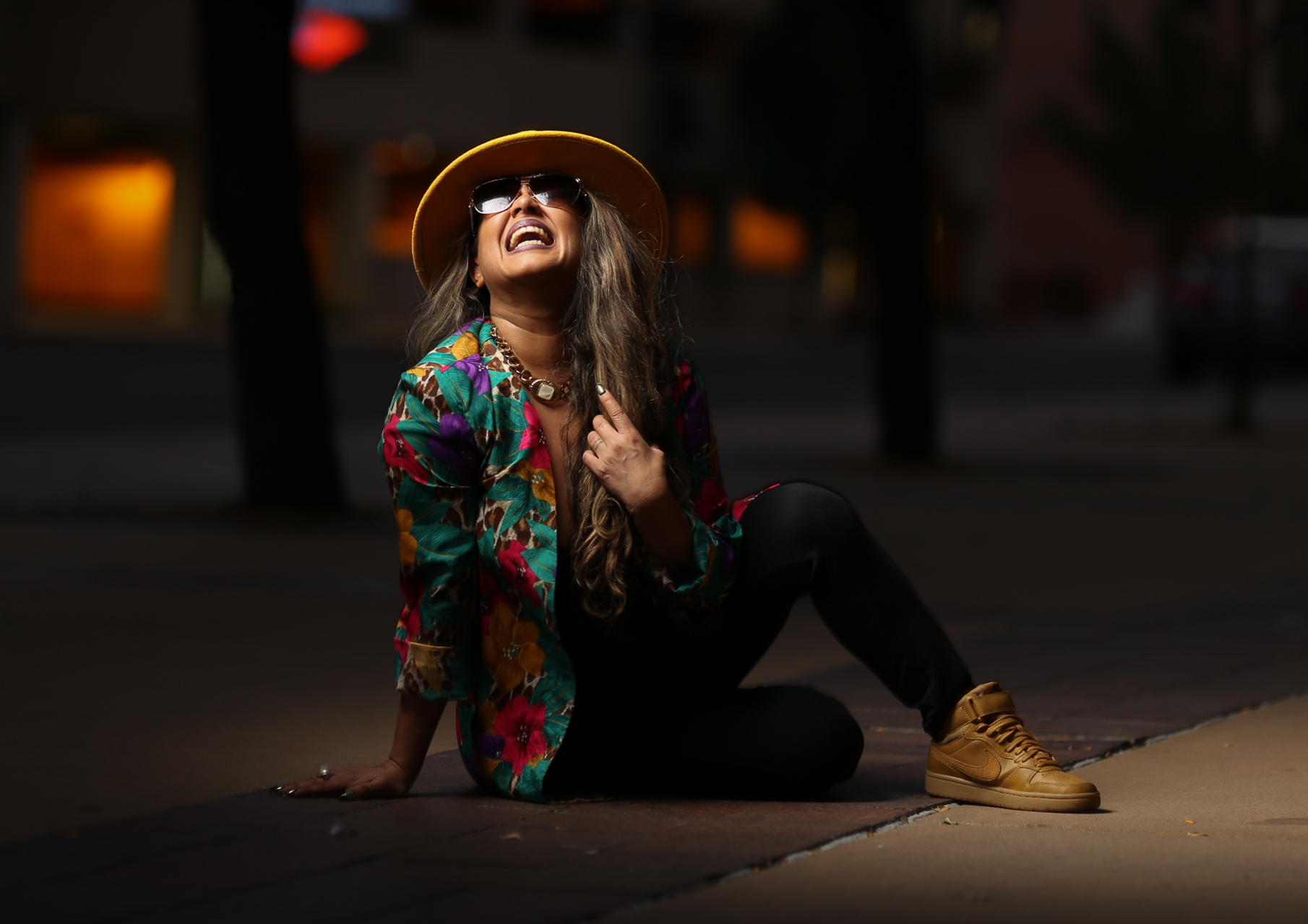 A person with long hair wearing a hat and sunglasses poses on the floor