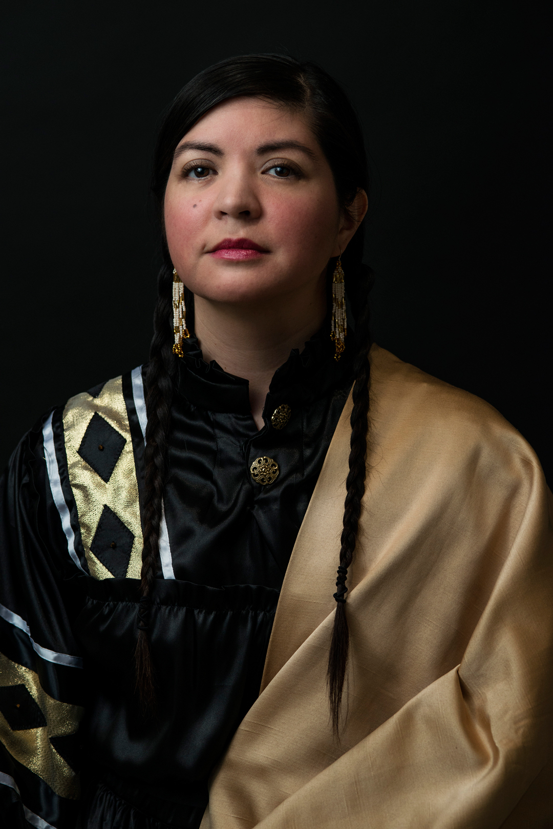 A woman dressed in black and gold Indigenous regalia with a long braid and red lip sits against a black background looking at the camera