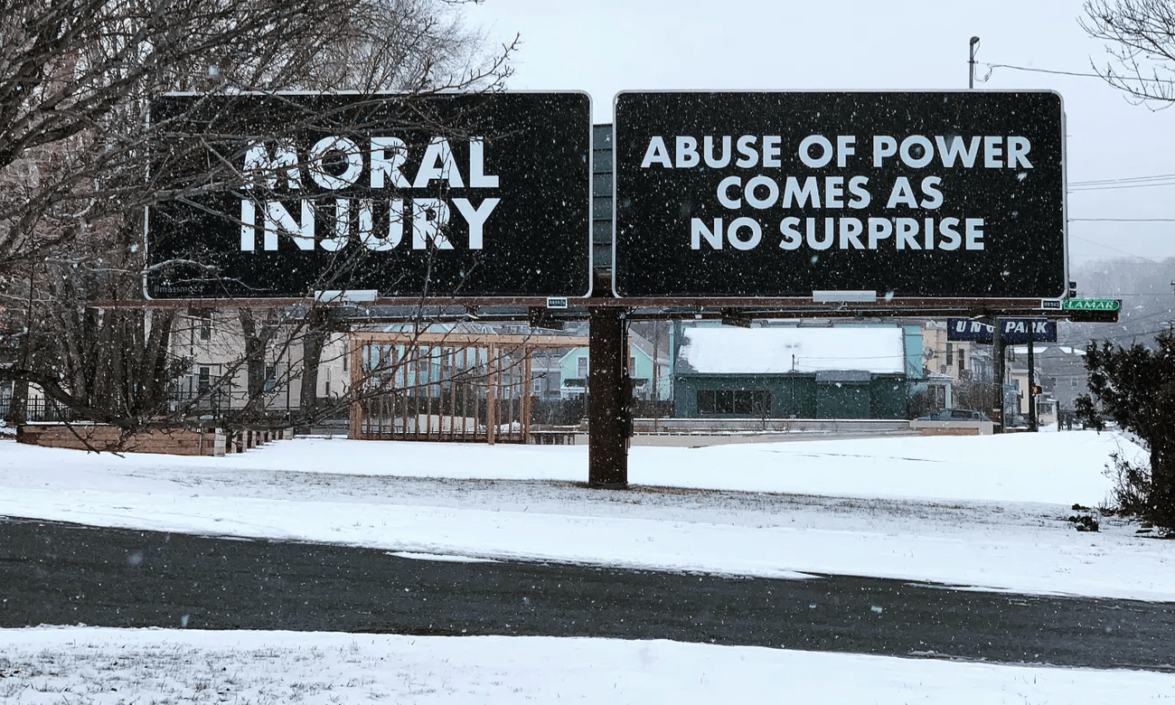 Two signs stand in the air surrounded by snow. The left reads MORAL INJURY. The right reads ABUSE OF POWER COMES AS NO SURPISE. They are in white letters on a black background.